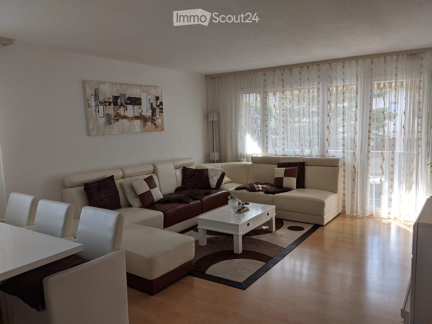Modern living room with a sectional sofa, coffee table, dining chairs, and artwork on the wall. The room features large windows with sheer curtains and a wooden floor.