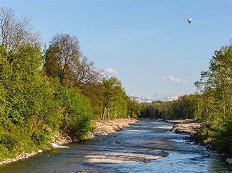 A serene river scene surrounded by lush green trees and a clear blue sky, with a distant view of mountains and a hot air balloon in the sky.