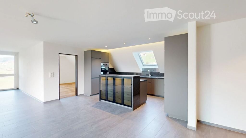 Open-plan kitchen and living area with modern cabinetry, a central island, and large windows. The space is unfurnished, showing only the built-in kitchen units and flooring.