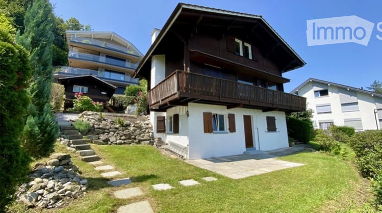 A two-story wooden house with a balcony, surrounded by greenery and a stone pathway leading to the entrance.
