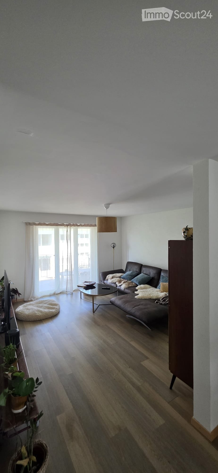 Modern living room with wooden flooring, large windows with white curtains, a dark brown leather sofa, a glass coffee table, a floor lamp, and potted plants.