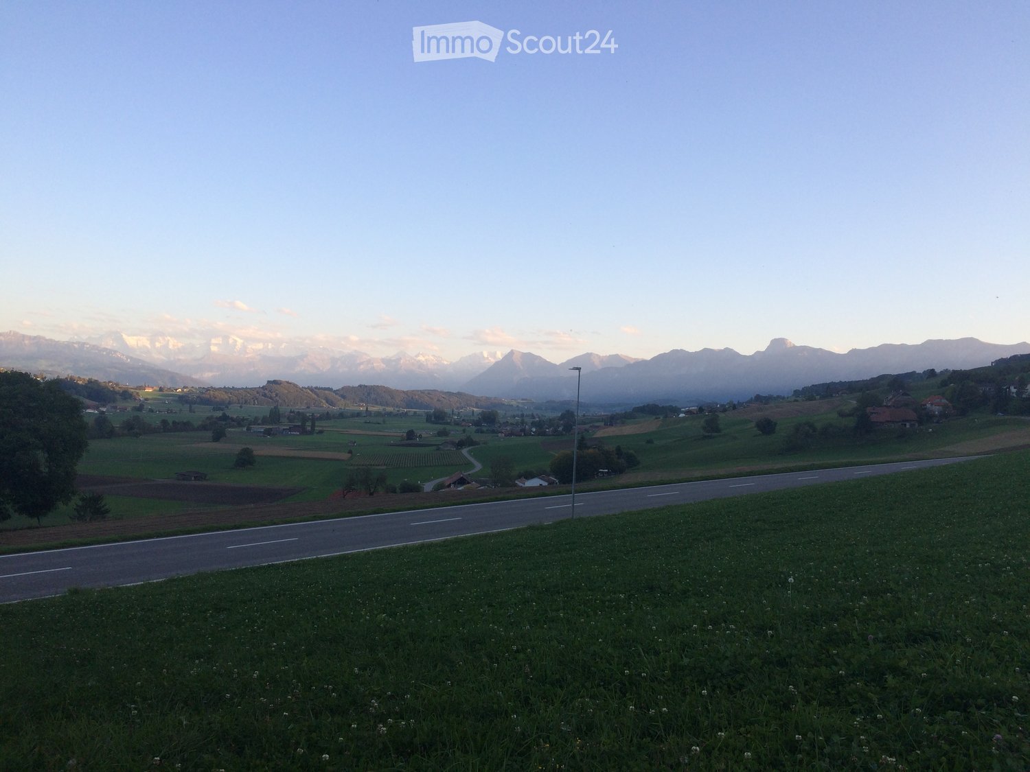 A peaceful rural landscape with green fields, scattered houses, and distant snow-capped mountains under a clear sky.