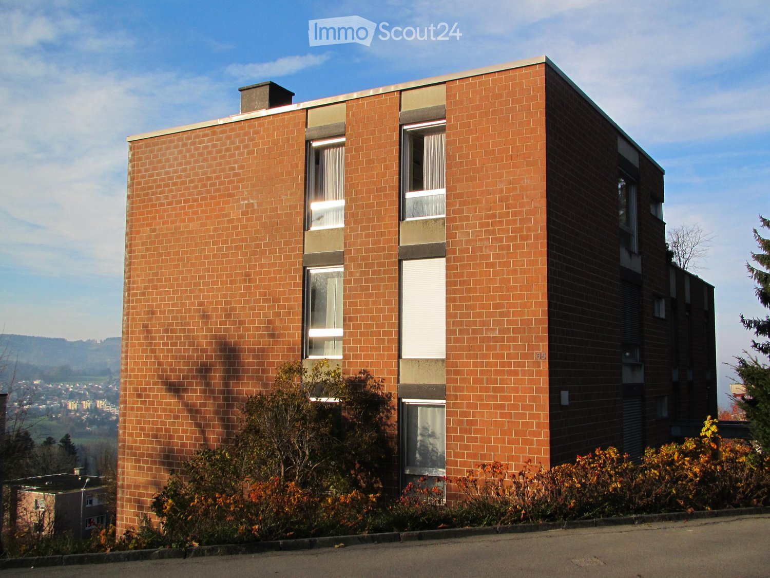 Modern two-story brick building with large windows and a flat roof, surrounded by greenery and shrubs.