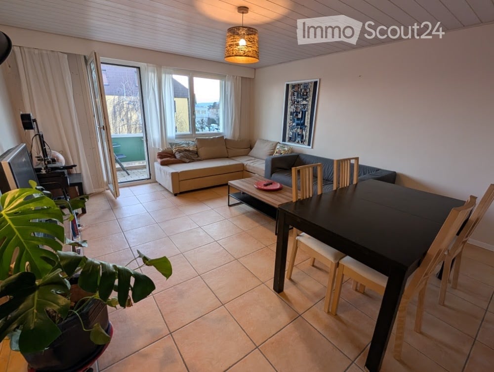 Open-plan living and dining area with a sectional sofa, coffee table, dining table with chairs, and a potted plant near the entrance to a balcony.