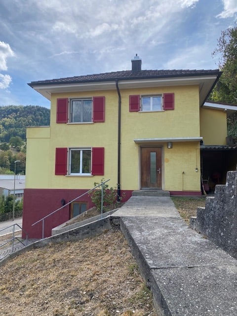 Two-story house with yellow and red exterior, red shutters, and a raised ground floor accessed by concrete steps.