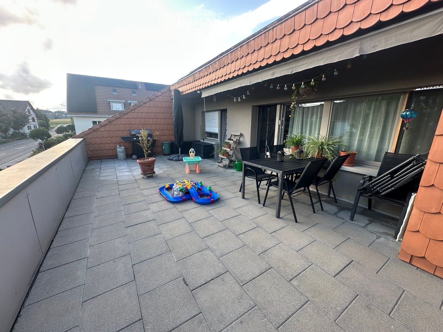 Balcony with tiled floor, outdoor furniture including a table and chairs, potted plants, and children's toys.
