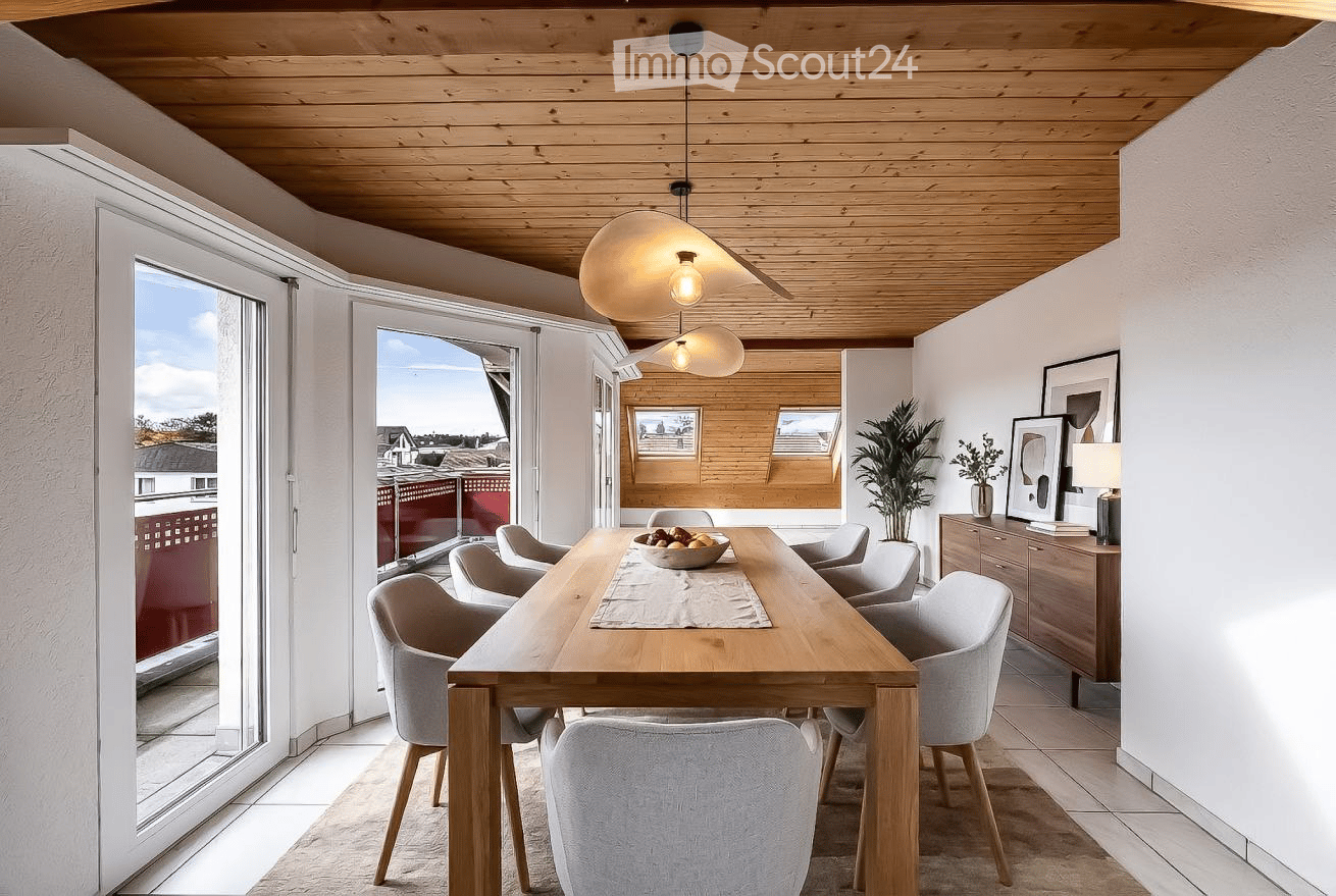 Bright dining area with a wooden table, six chairs, a bowl of fruit, a cabinet with a lamp, and a view of the city through large windows.