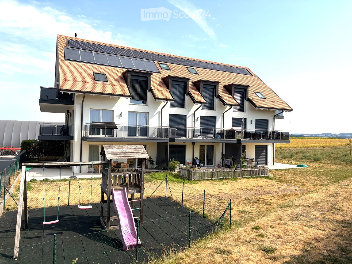 Modern two-story house with solar panels on the roof, large windows, balconies, and a fenced yard with a playground featuring a swing and slide.