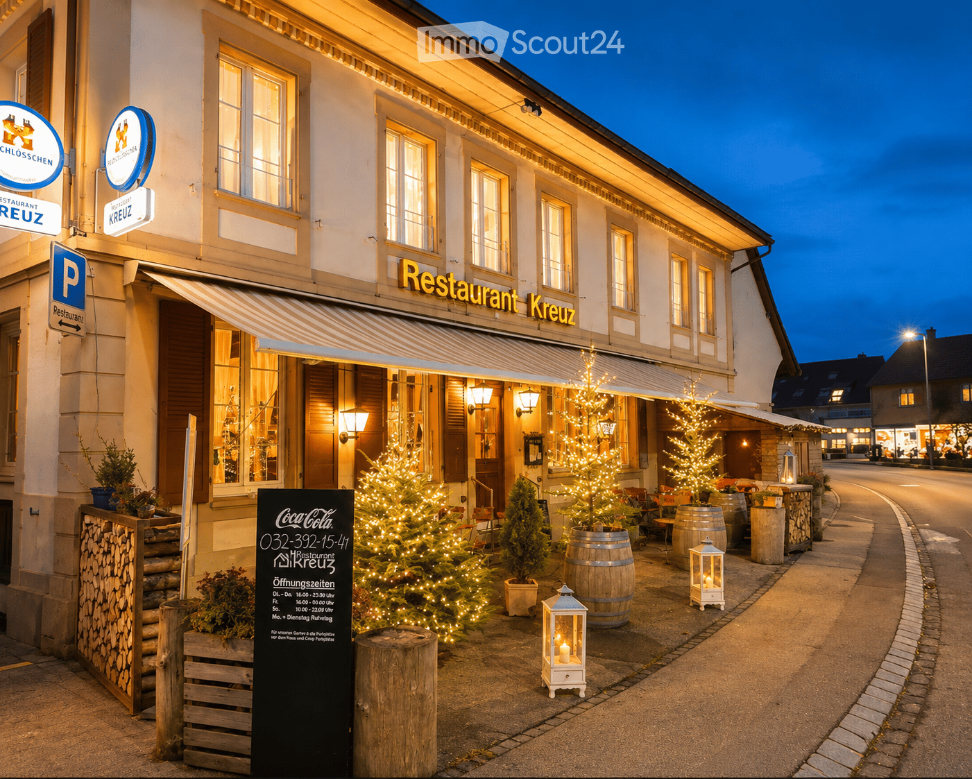 Exterior view of Restaurant Kreuz at dusk, featuring illuminated windows, festive Christmas trees, wooden barrels, and a signboard with operating hours.