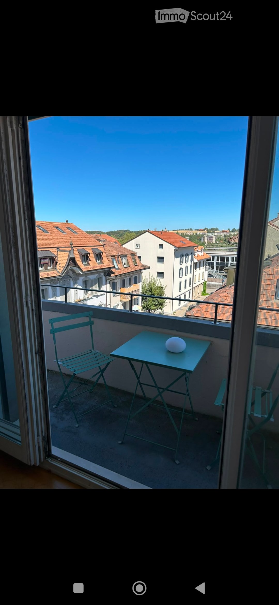 Balcony with a small table and a chair, overlooking a cityscape with red-tiled roofs and buildings.