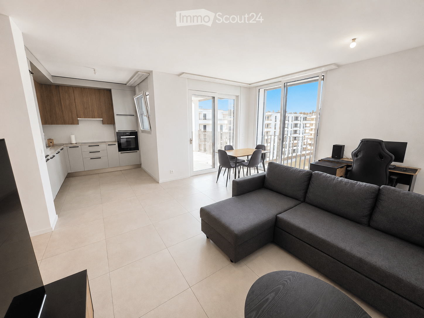 Modern, open-plan living and dining area with a grey sectional sofa, ottoman, dining table with chairs, and a kitchen area with cabinets and an oven. A desk with a computer setup is also visible.