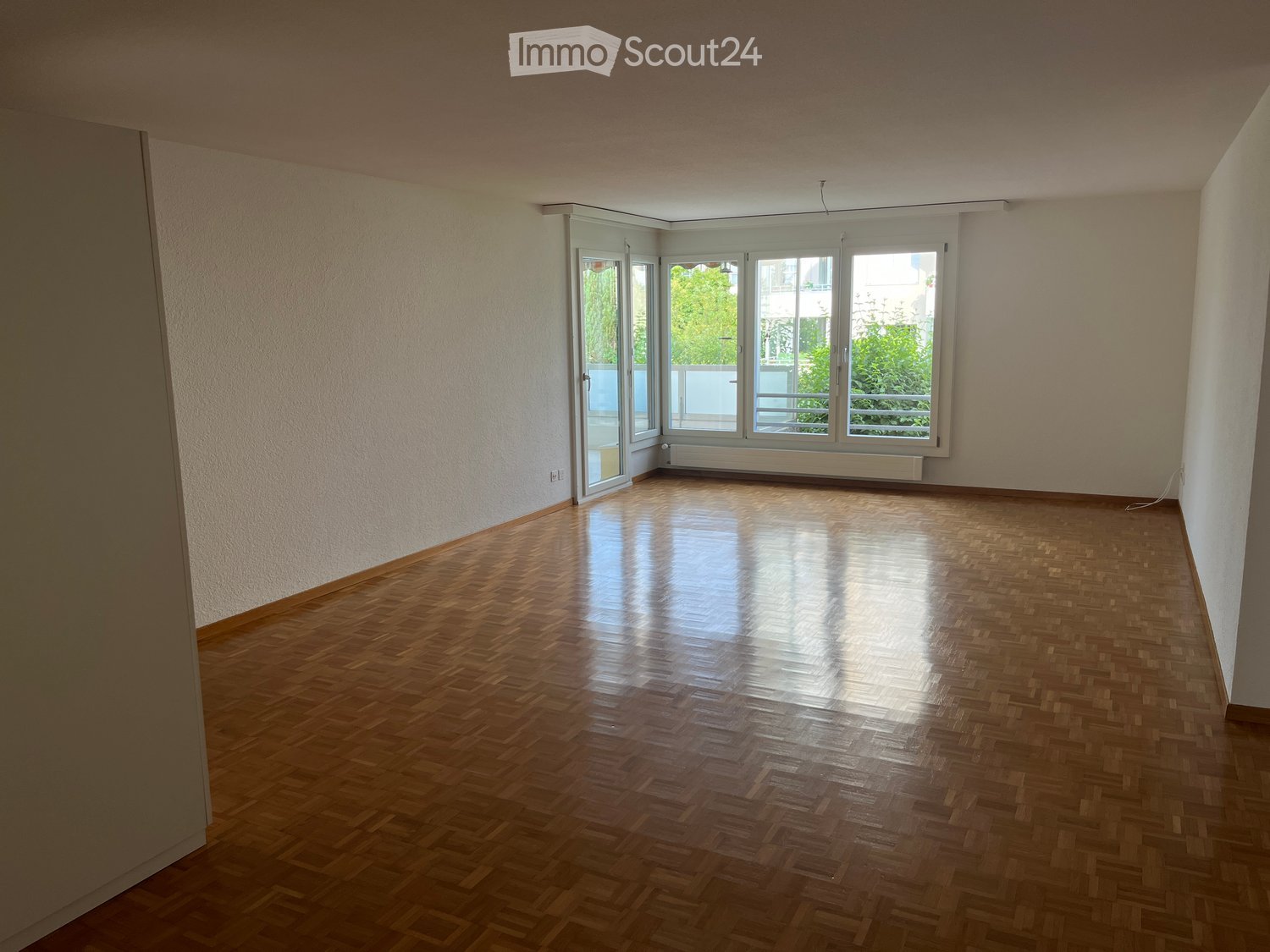 Empty room with wooden parquet flooring, large windows, and white walls.