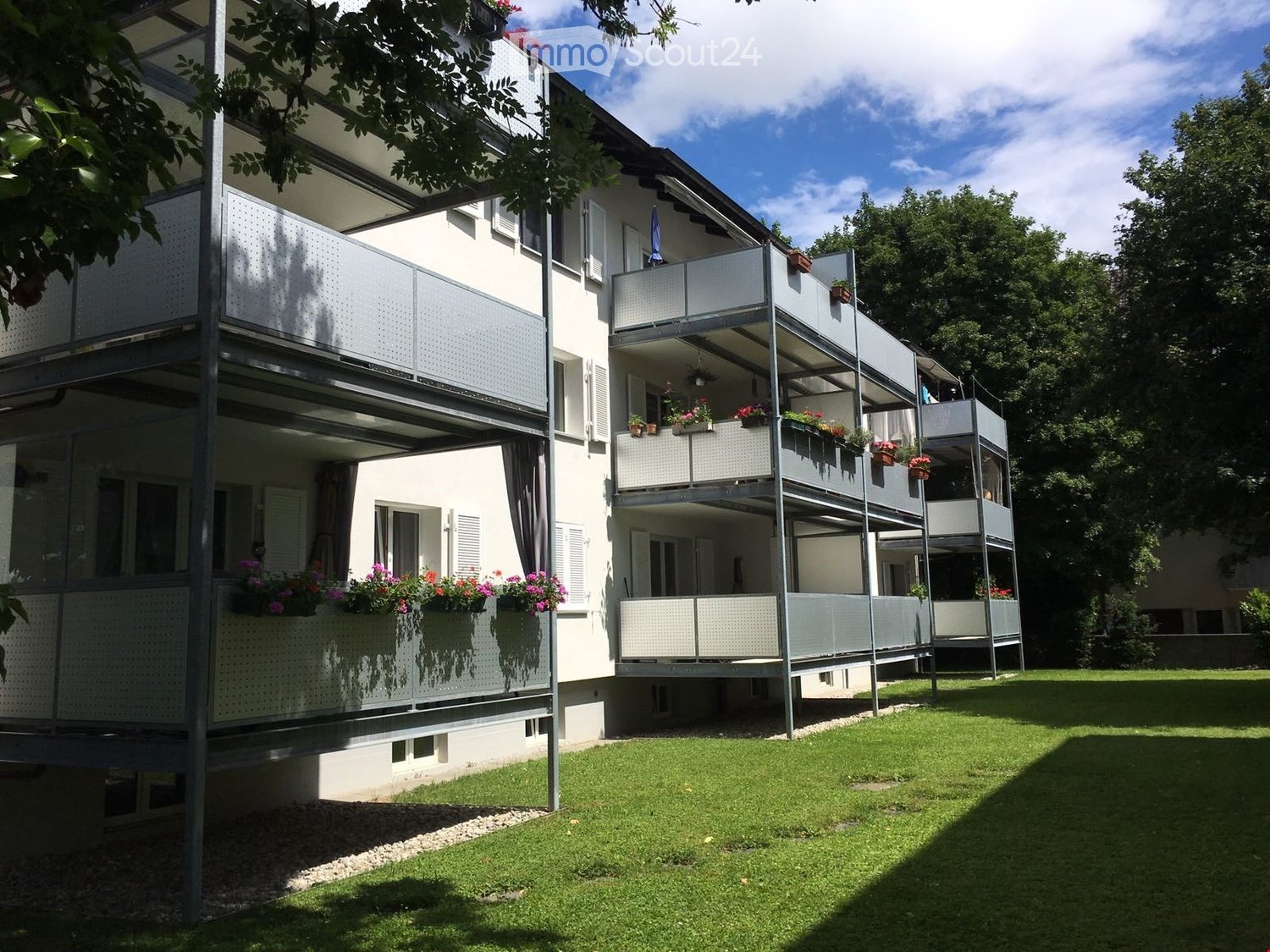 Multi-story residential building with balconies, white exterior, and flower boxes on the balconies. The building is surrounded by a well-maintained lawn and trees.
