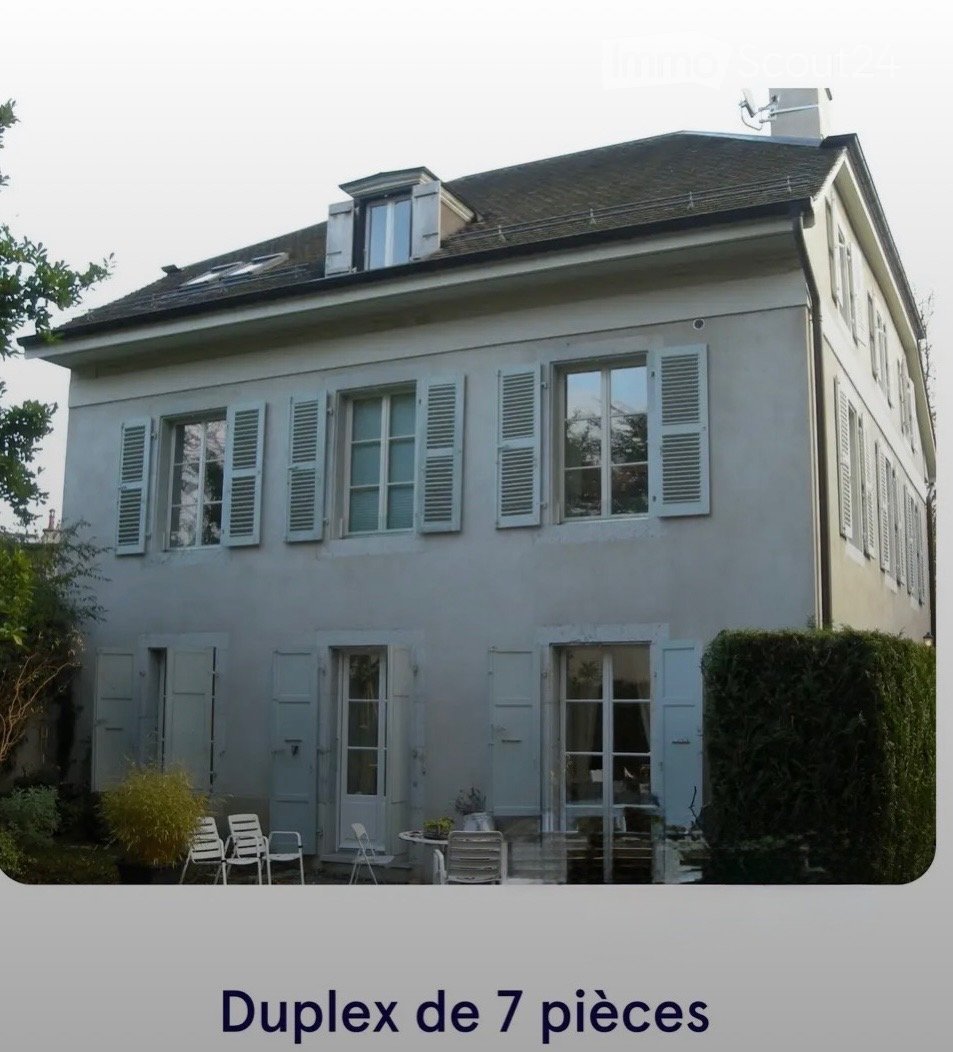 Two-story house with light gray facade, multiple windows with shutters, and a small outdoor seating area with chairs and a table.