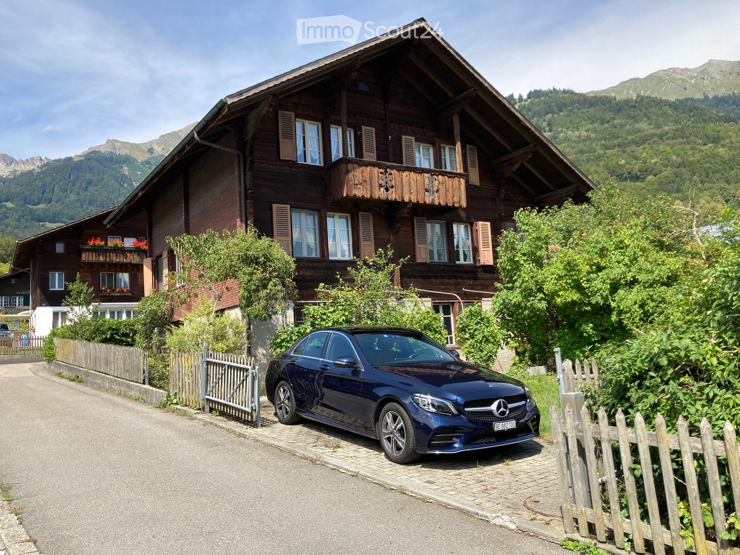A two-story wooden house with white-framed windows, balconies, and a blue Mercedes-Benz parked in front.