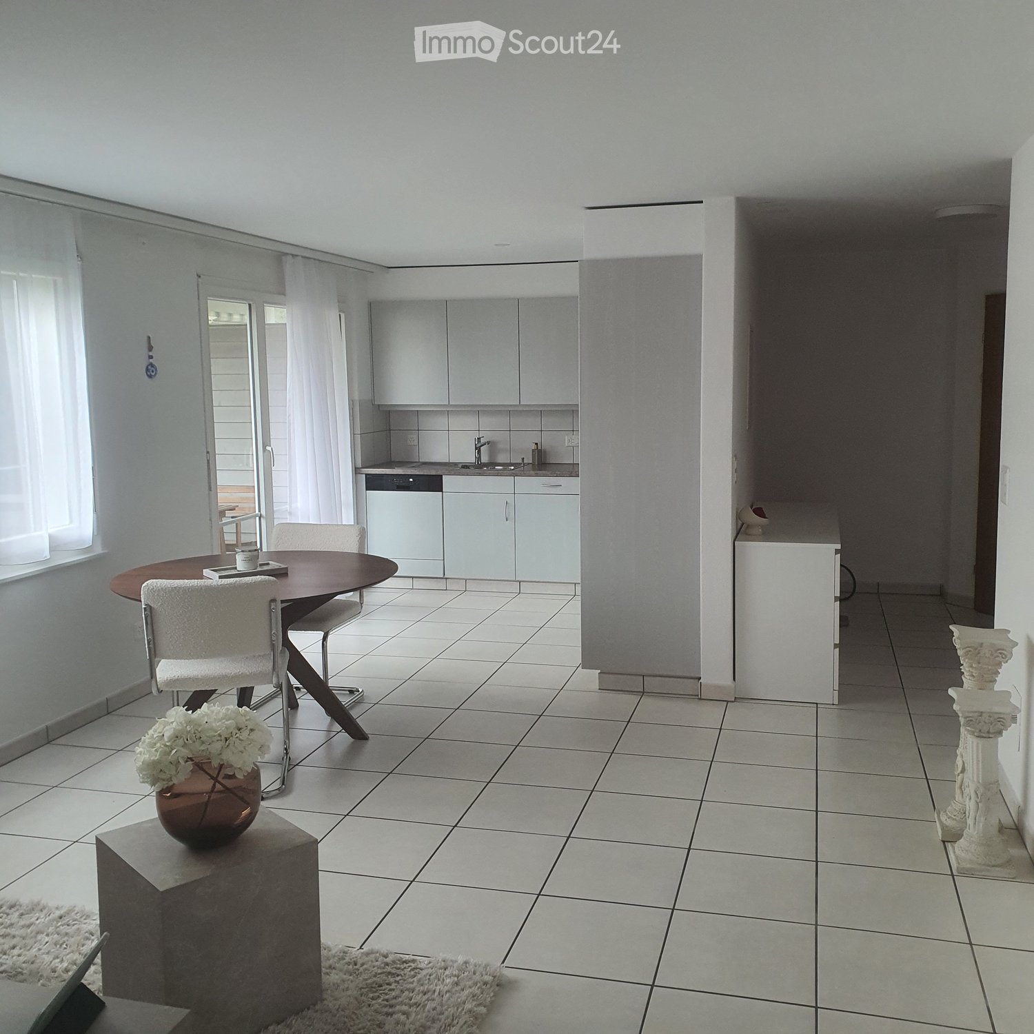 Bright, modern room with tiled flooring, a round wooden dining table with four white chairs, a vase with white flowers on a small table, and a kitchen area with white cabinets and a sink.