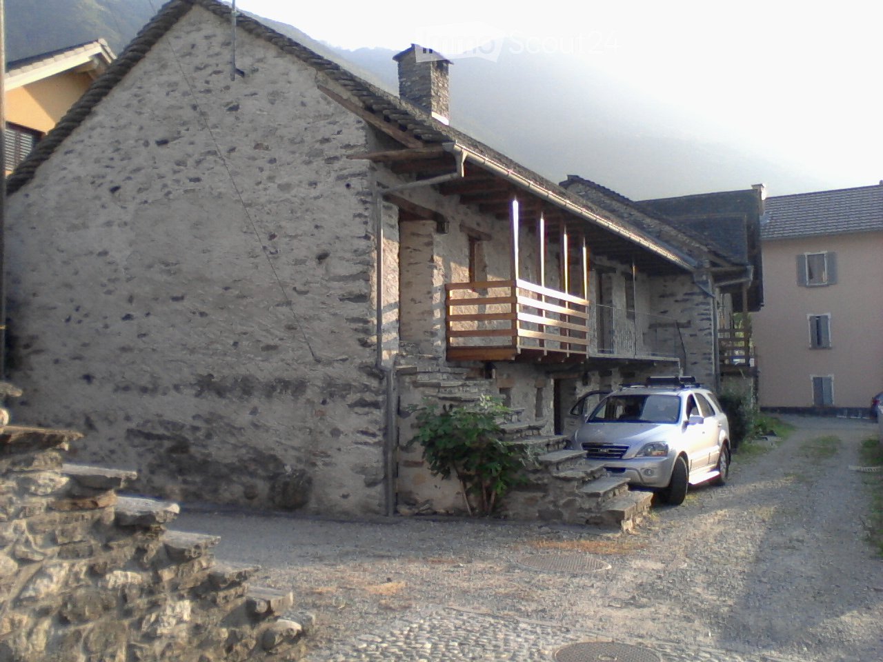 A rustic stone house with a wooden balcony and a parked car in front.