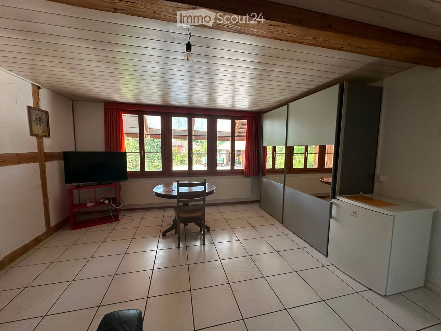 A furnished room with tiled flooring, a round wooden table with a single chair, a television on a red stand, a refrigerator, and a cabinet. The room has large windows with red curtains and a slanted wooden ceiling.