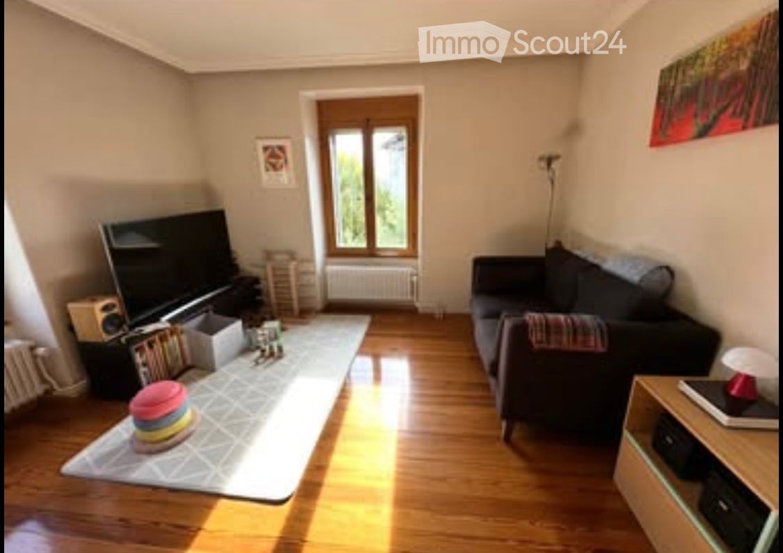 A cozy living room with wooden flooring, a large window, a black sofa, a coffee table, a television, and a speaker system.