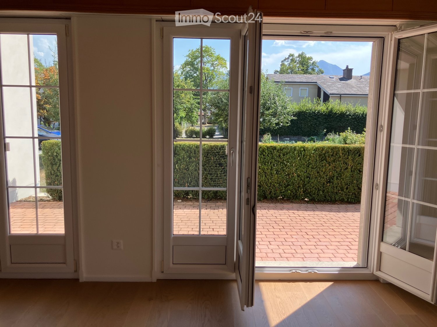 Empty room with wooden flooring, large open sliding doors leading to a brick patio and garden area.