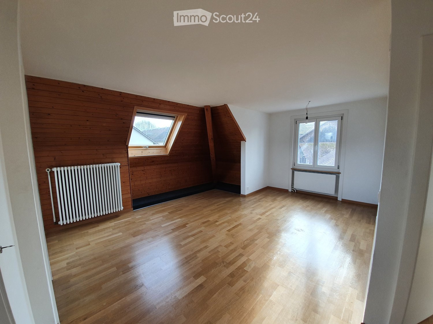 Empty room with wooden flooring, white walls, and two windows, one of which is a skylight. Radiators are present on both sides of the room.