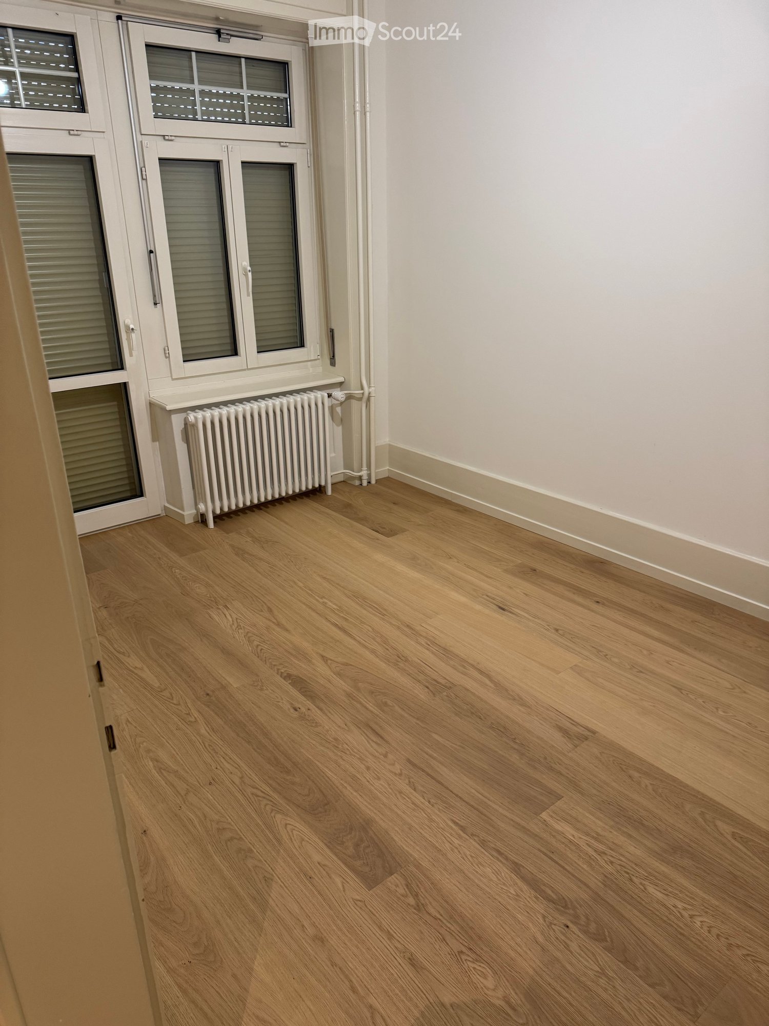 Empty room with light wooden flooring, white walls, and a radiator near a window with blinds.