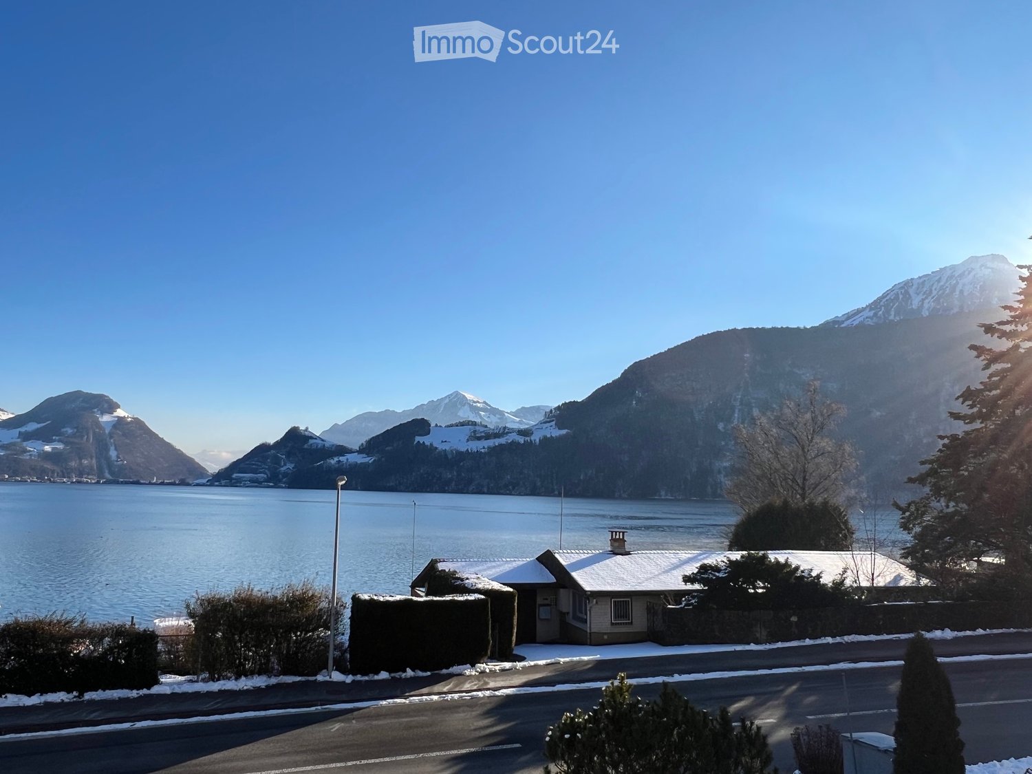 A small house with a snow-covered roof is situated near a body of water, with snow-capped mountains in the background.