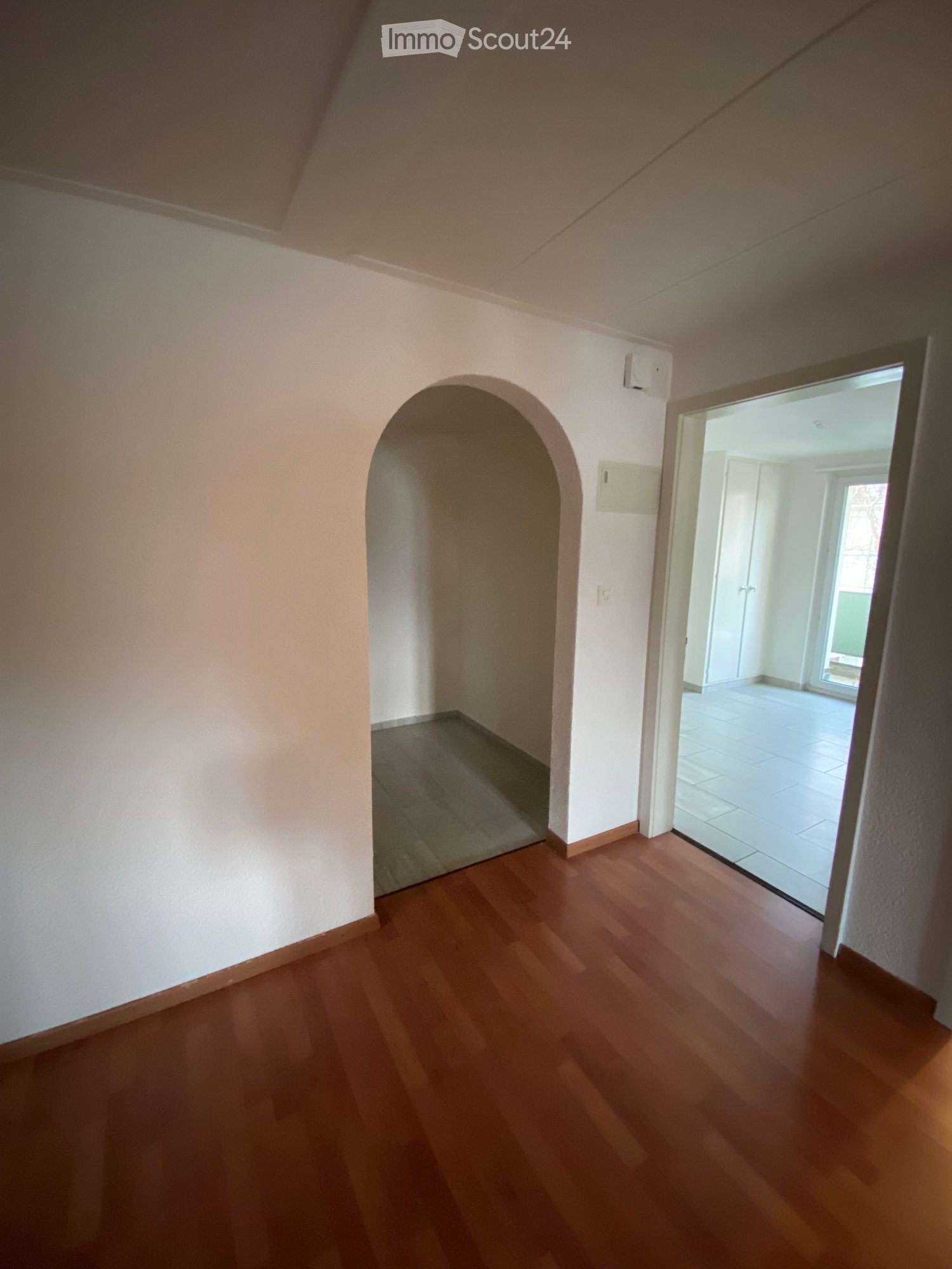 Empty hallway with wooden flooring, white walls, and an arched doorway leading to another room with tiled flooring.