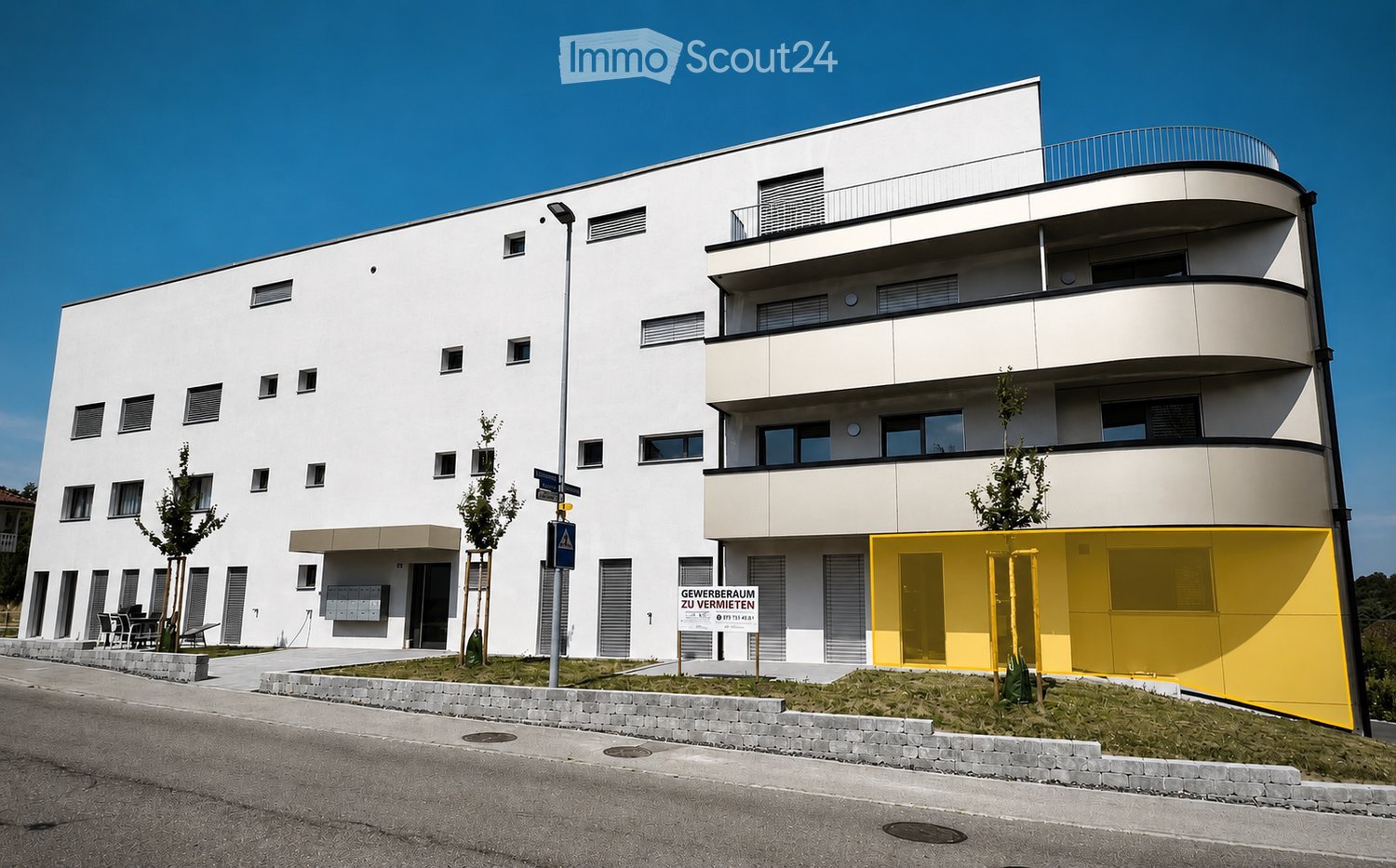 Modern two-story building with white facade, multiple windows, and a curved balcony section on the upper floor. A yellow garage door is visible on the right side.