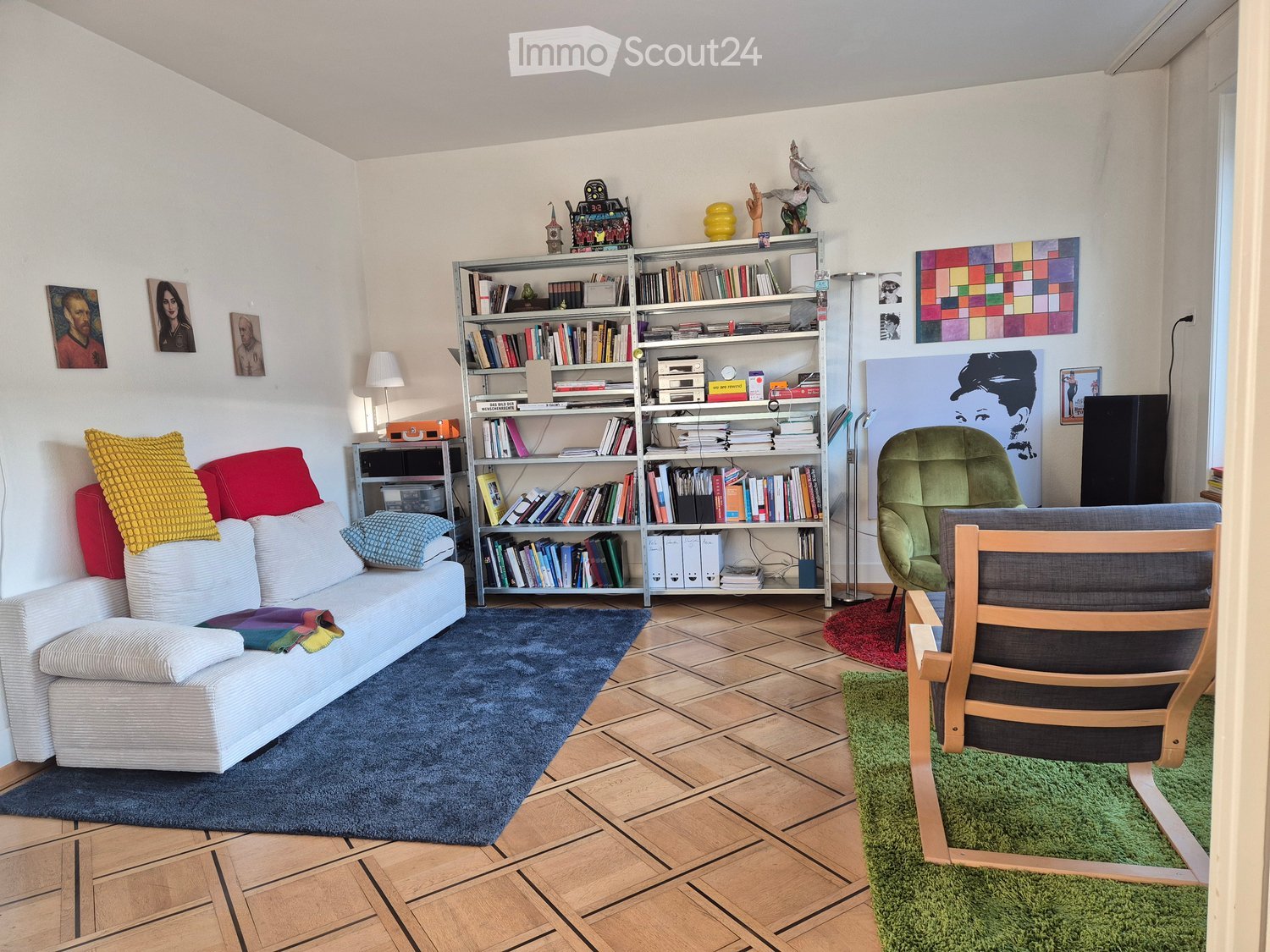 Living room with a white sofa, colorful cushions, a bookshelf filled with books, a green armchair, a wooden chair, and various decorative items on shelves and walls.