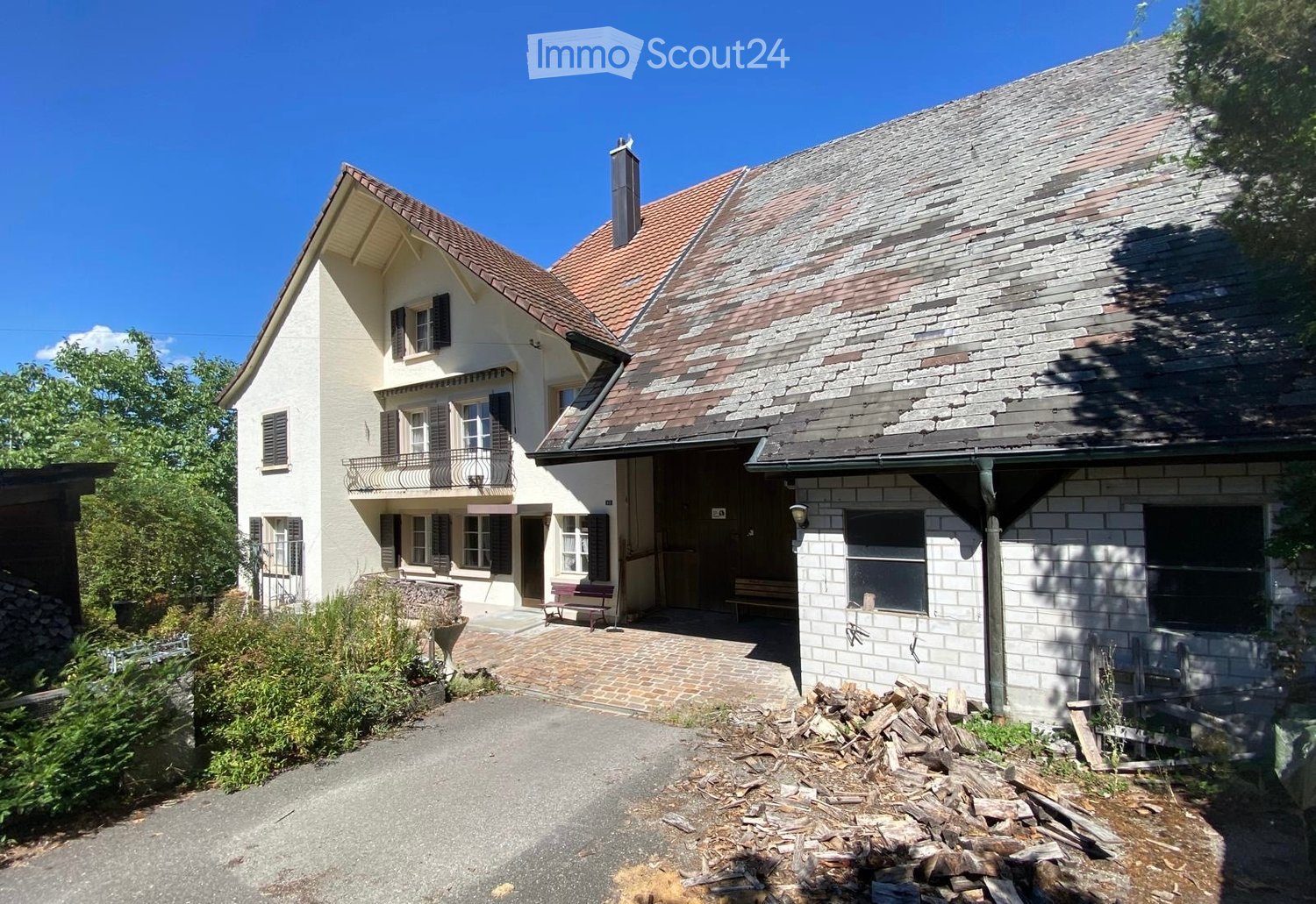 Two-story house with a red-tiled roof, white walls, multiple windows with shutters, and a small balcony. The house has a stone chimney and a brick pathway leading to the entrance. There is a stack of firewood and some greenery around the house.