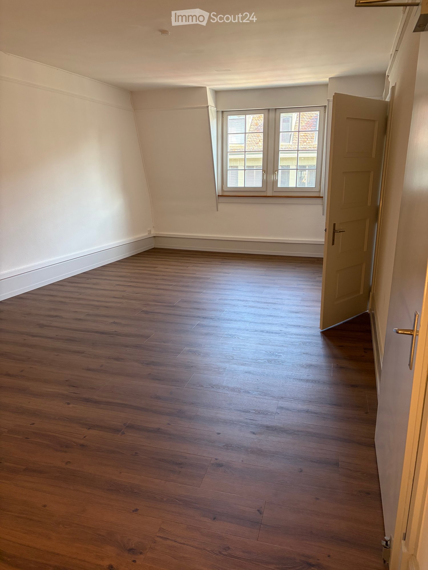 Empty room with wooden flooring, white walls, and a window with a view of rooftops.