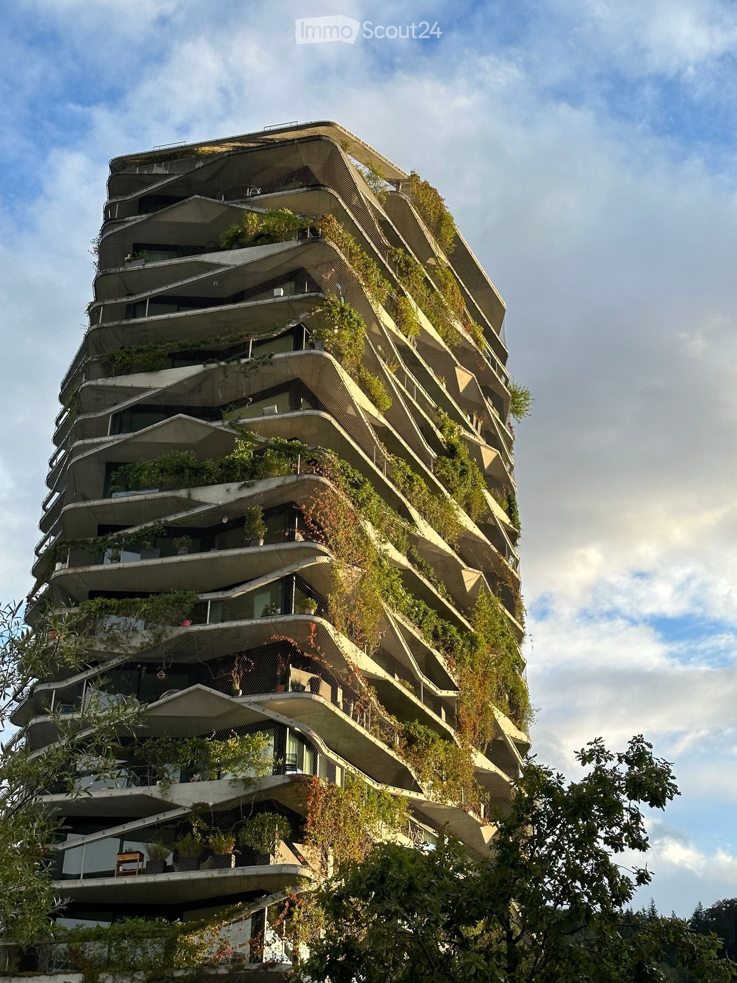A modern, multi-story building with a green facade covered in plants and trees, featuring numerous balconies and terraces.