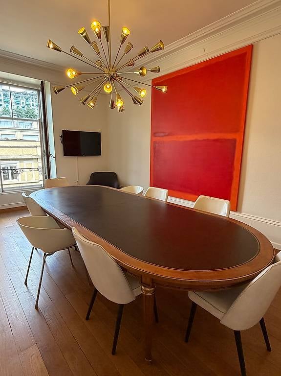 A modern, well-lit room with a large, curved wooden table surrounded by white chairs, a television mounted on the wall, and a colorful abstract painting. A large, multi-bulb chandelier hangs from the ceiling, and large windows offer a view of the city.