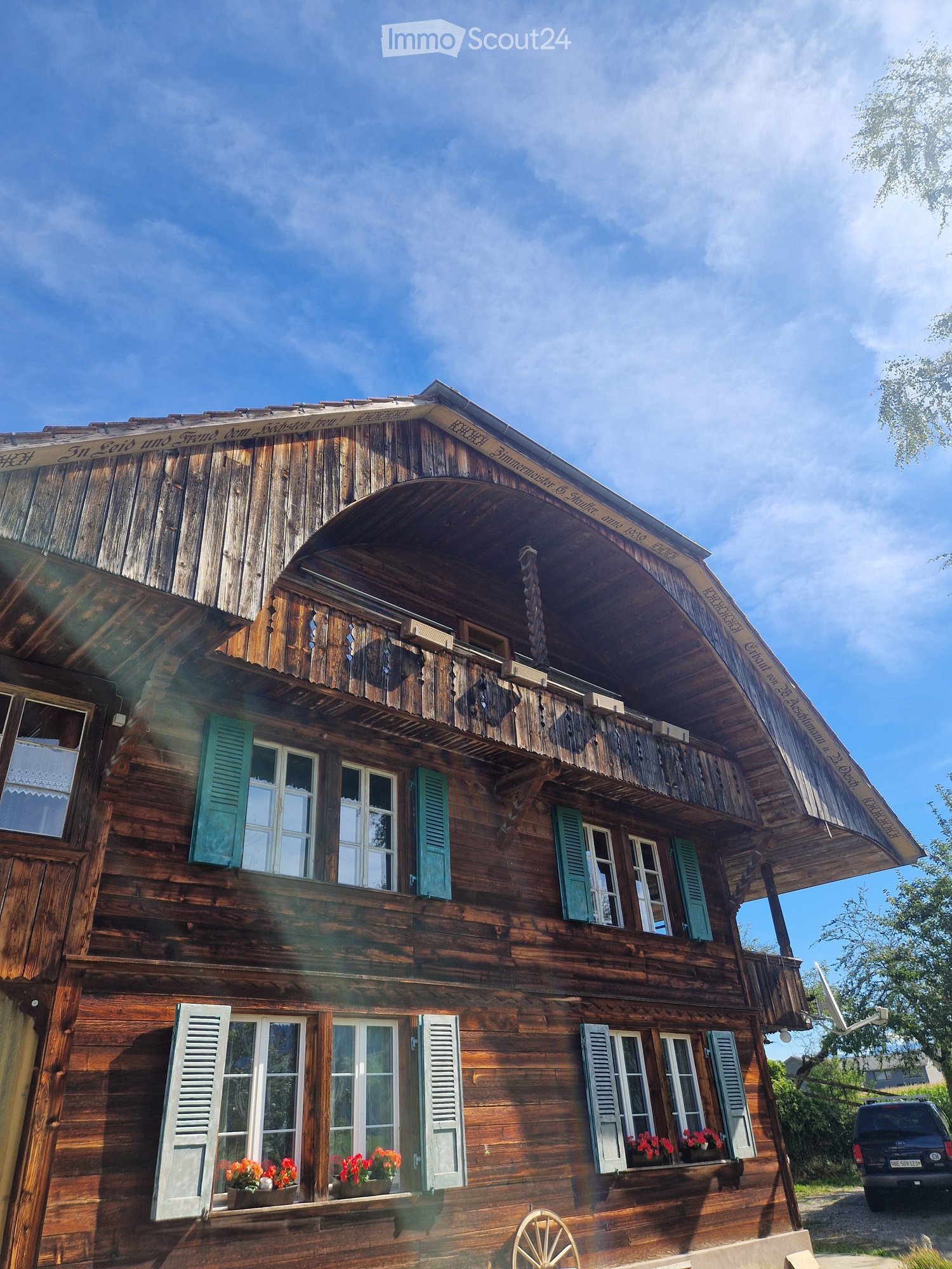 Two-story wooden house with green shutters, white-framed windows, and flower boxes on the lower level. The roof has a decorative wooden overhang and a balcony on the upper level.