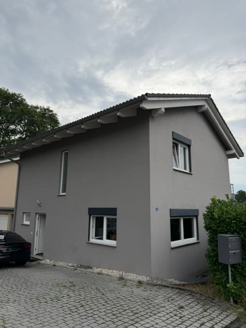 Single-story house with grey exterior, white-framed windows, and a small driveway.