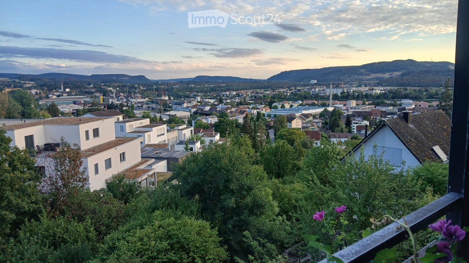A balcony view overlooking a town with houses, trees, and distant hills under a partly cloudy sky.