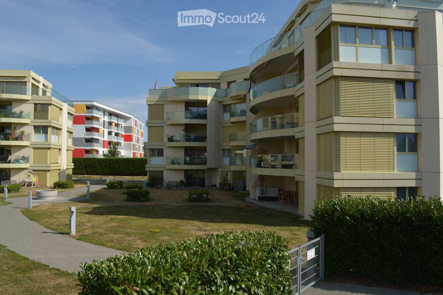Modern multi-story residential building with balconies, glass railings, and a landscaped courtyard area with pathways and greenery.