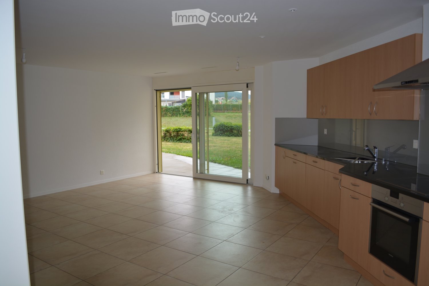 Modern kitchen with light wooden cabinets, black countertops, and stainless steel appliances. Features a large sliding glass door leading to a green outdoor area.