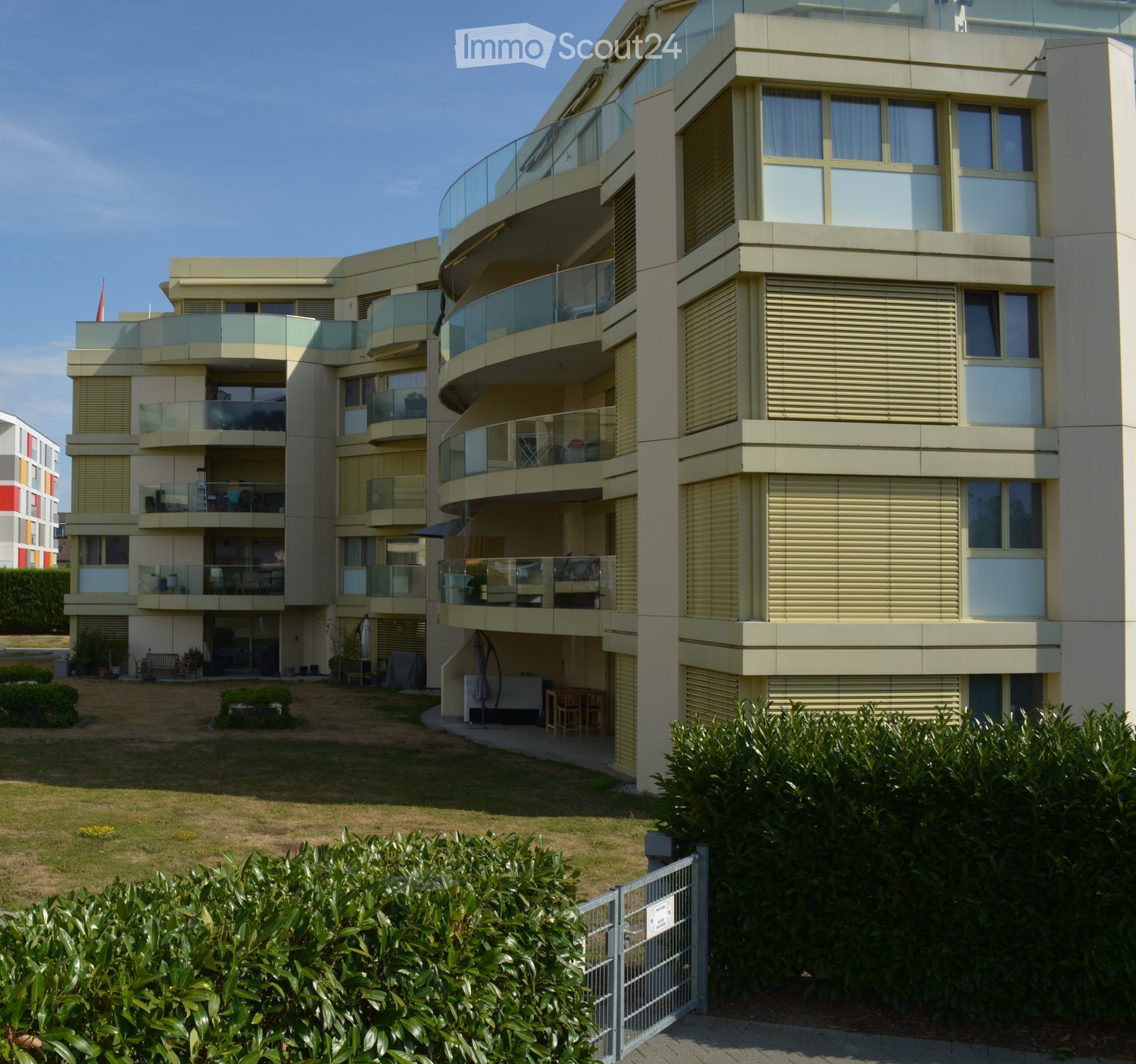Modern multi-story residential building with balconies, large windows, and a landscaped garden area.