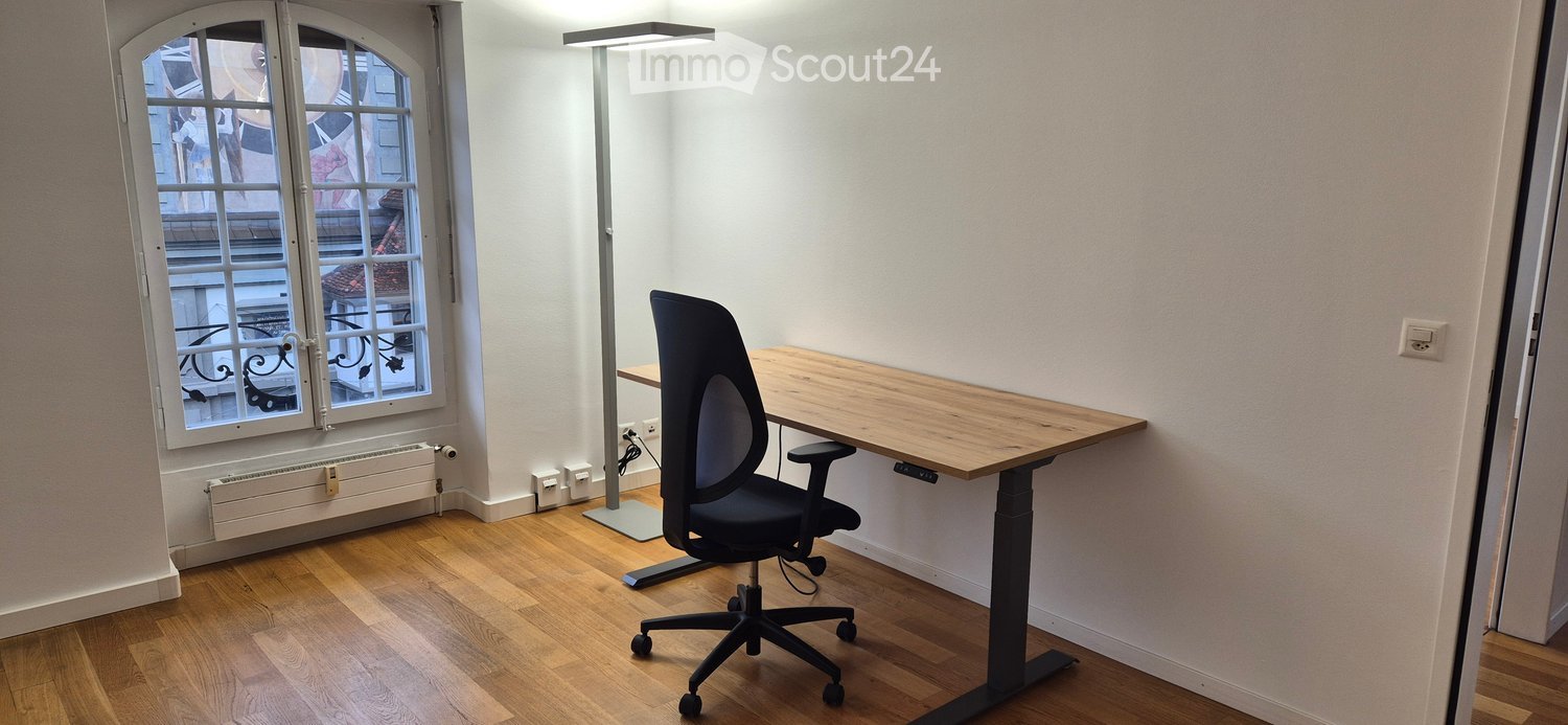 A modern, minimalist room with wooden flooring, a large window with decorative ironwork, a sleek desk, and a comfortable office chair.
