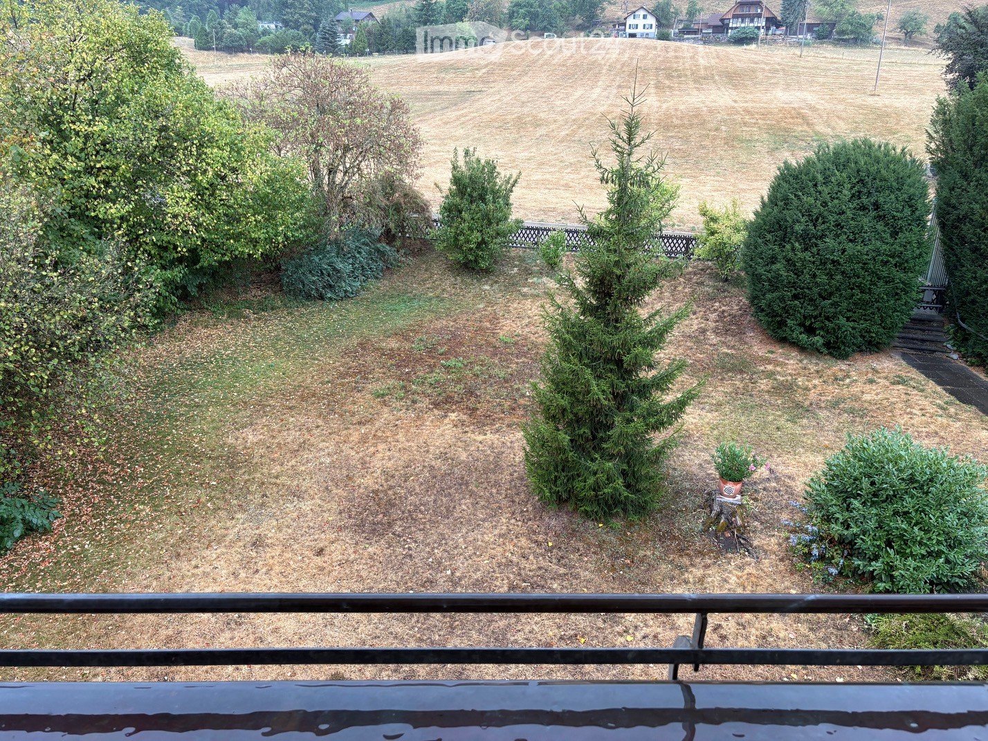 View of a garden area with grass, shrubs, and trees, overlooking a field and distant houses.