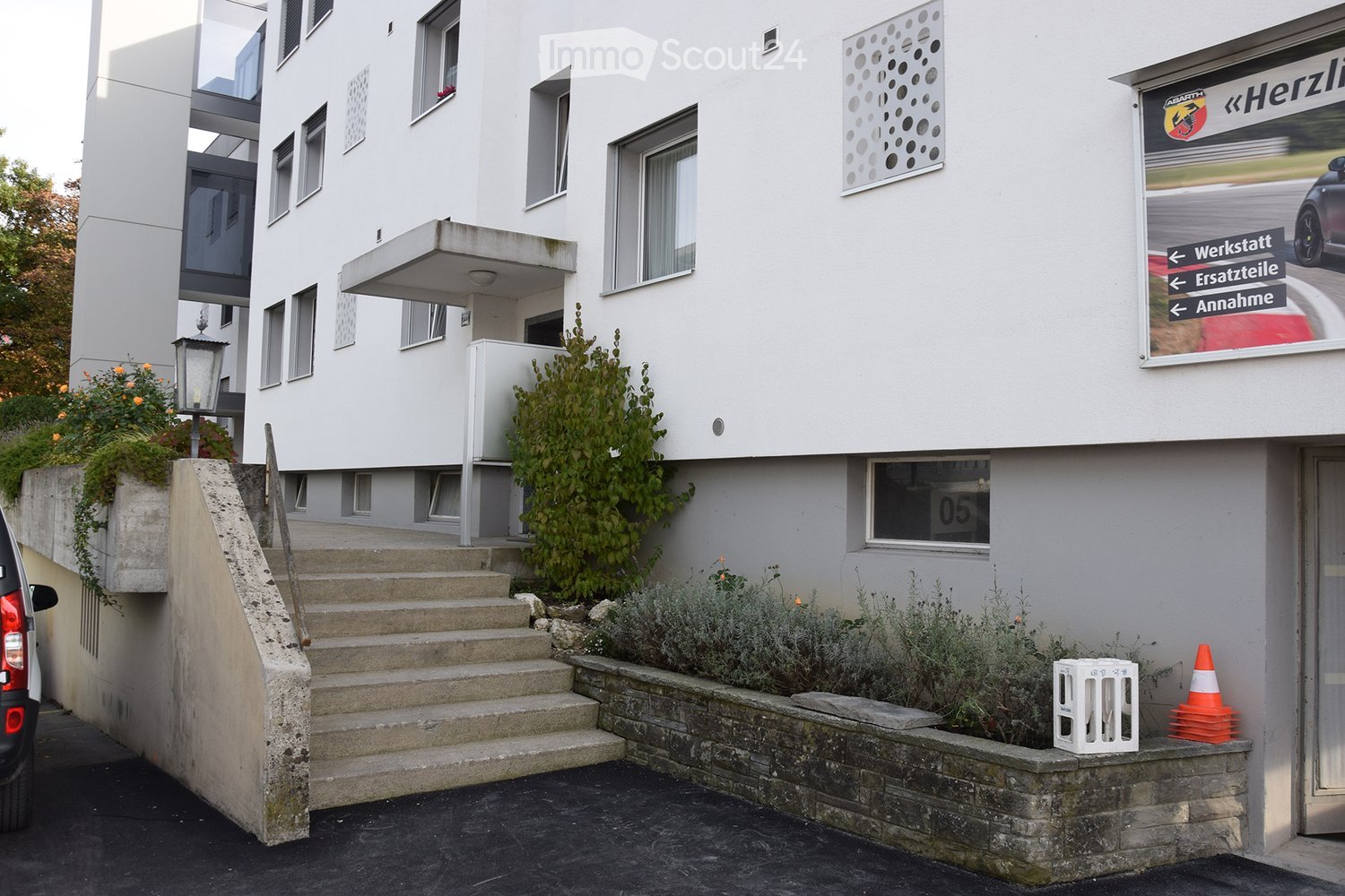 Modern two-story building with white facade, multiple windows, and a raised ground floor entrance accessed by concrete steps. The building features signage for a car service center, including directions to Werkstatt, Ersatzteile, and Annahme. There is a s