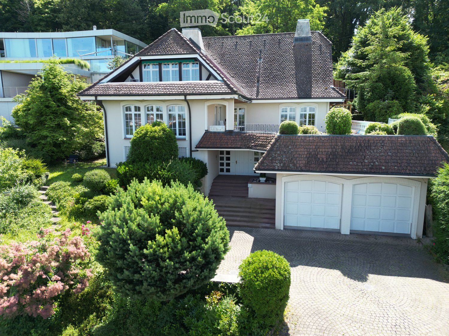 A two-story house with a tiled roof, white exterior, and a large garage. The property features a well-maintained garden with shrubs and a paved driveway.