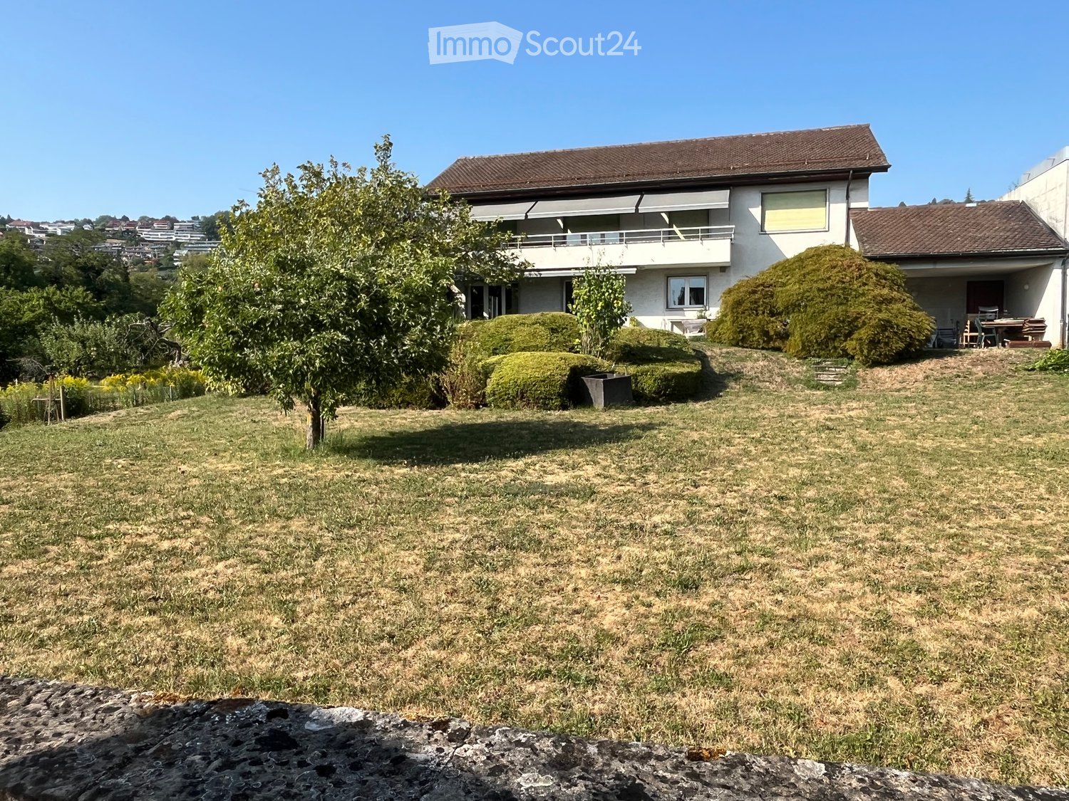 A well-maintained garden with a lawn, trees, shrubs, and a modern house with a balcony and patio furniture in the background.
