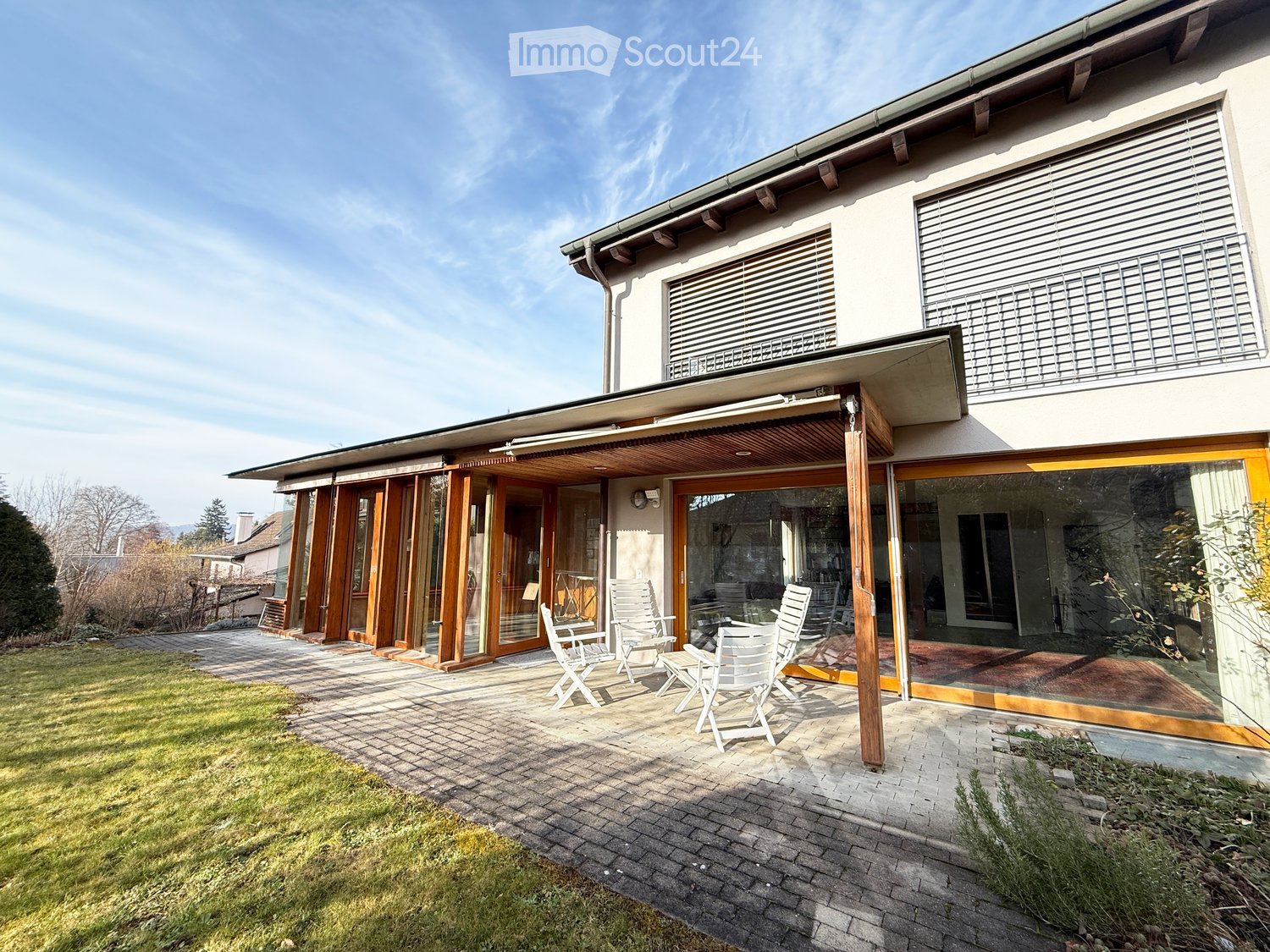 A modern house with a spacious outdoor patio area featuring white chairs and a table, surrounded by a well-maintained lawn and paved pathways.