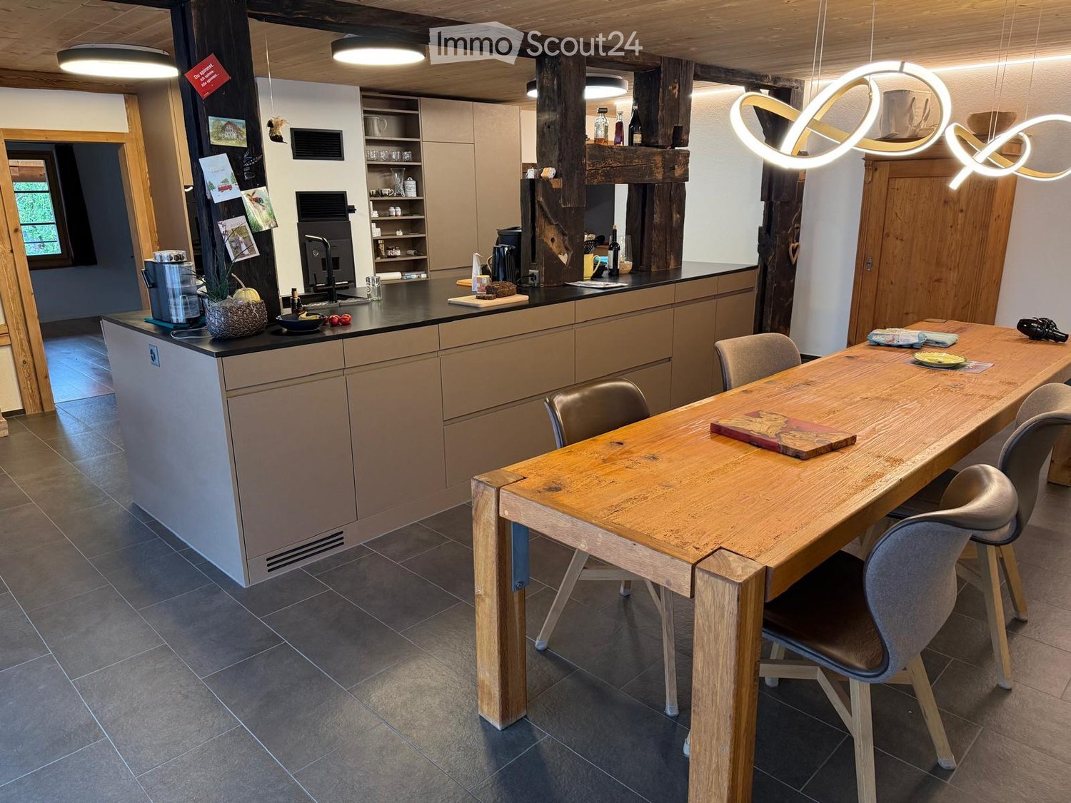 Modern kitchen with a wooden dining table, chairs, and various appliances including a refrigerator, sink, and coffee maker. The space features exposed wooden beams and a sleek, contemporary design.