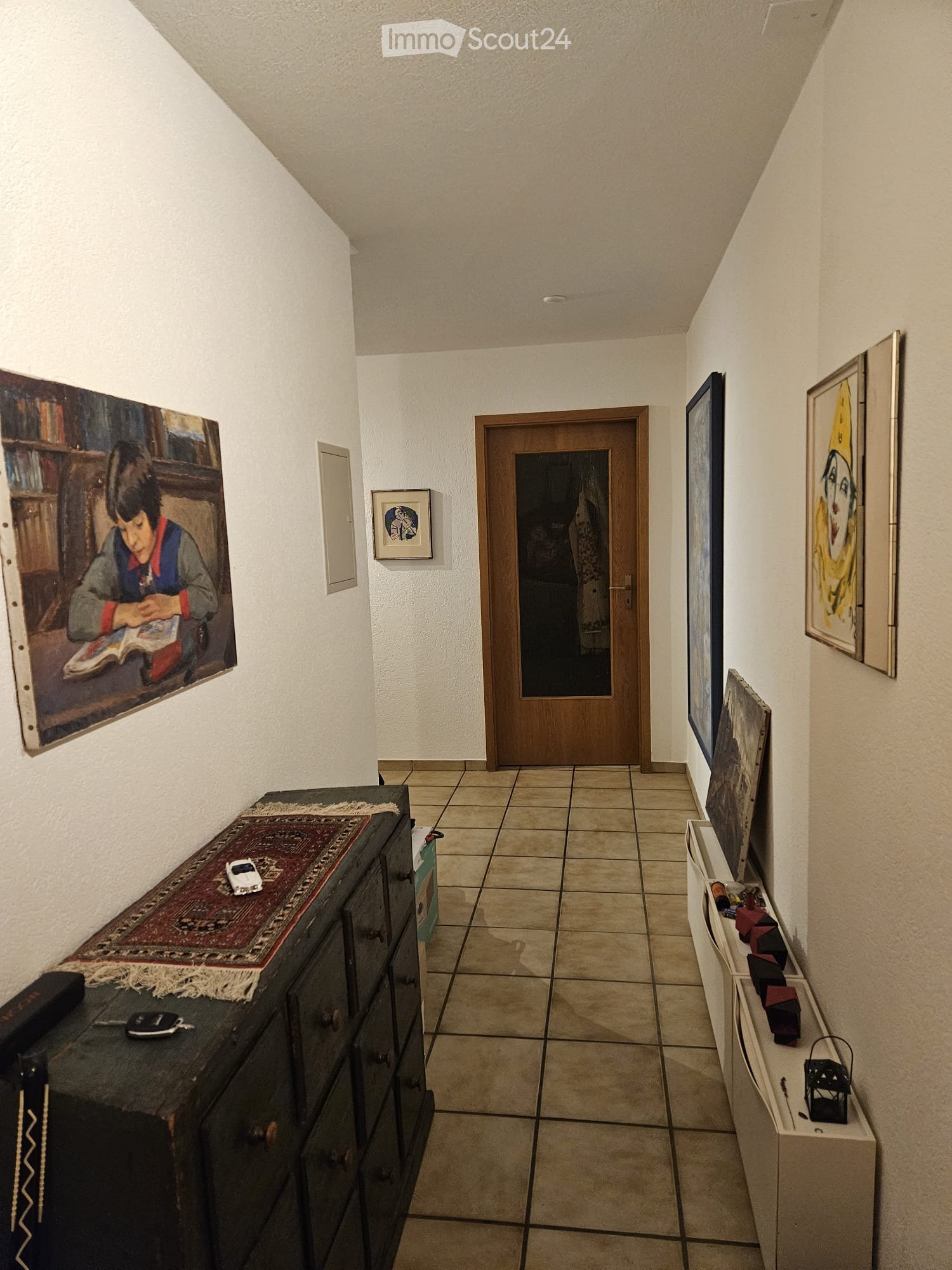A narrow hallway with tiled flooring, white walls, and several framed artworks displayed on the walls. A wooden door is visible at the end of the hallway, and a small table with a rug and some items is placed on the left side.