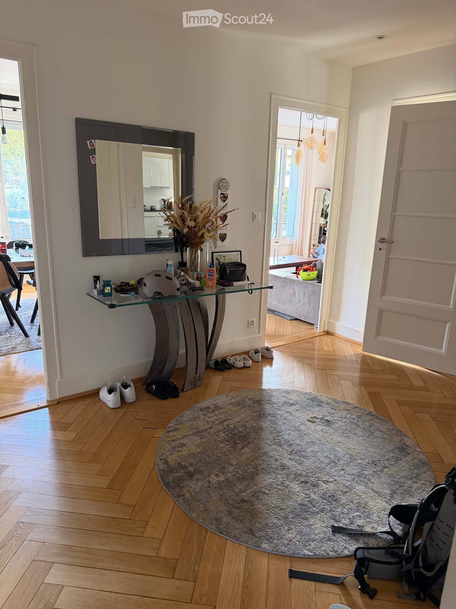 Modern hallway with wooden flooring, a round rug, a glass console table, and a mirror. Visible items include a chair, a small table, a vase with dried flowers, and a baby chair.