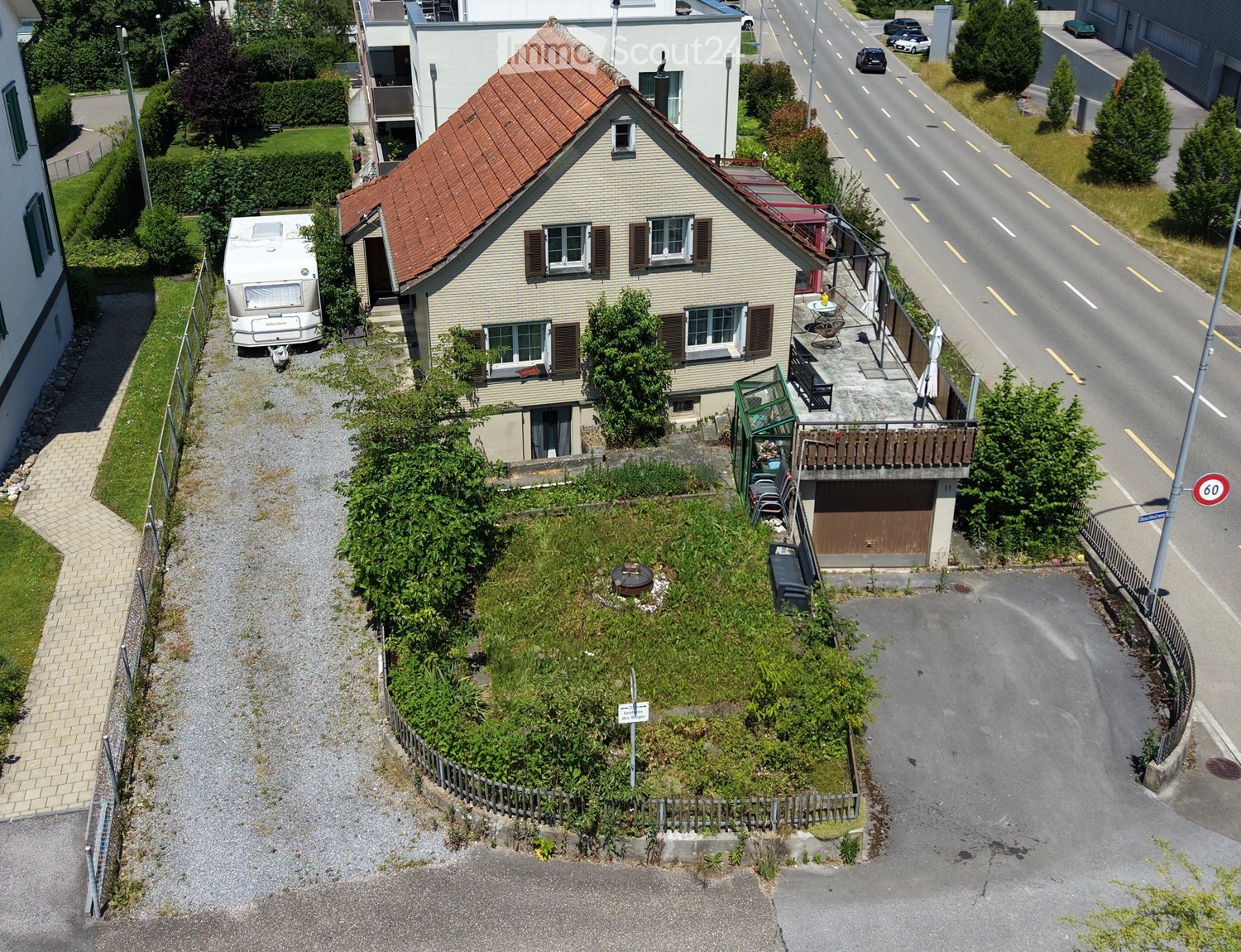 A two-story house with a red-tiled roof, multiple windows, and a small balcony area. There is a garage, a driveway, and a small garden area with a fence. A camper van is parked nearby.